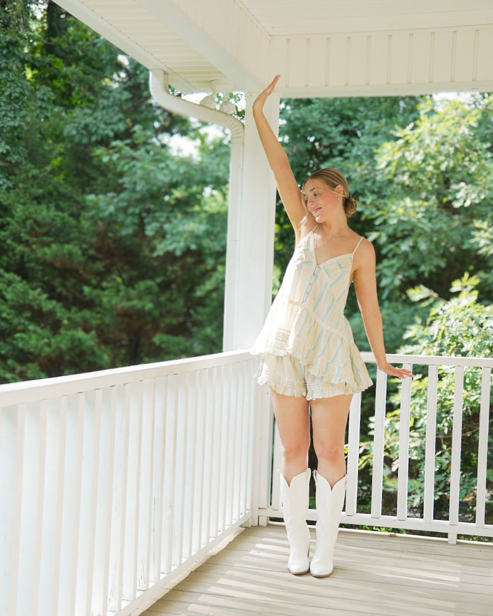 A woman stands on a white porch with trees in the background, wearing a light cream ruffled camisole and matching ruffled shorts with white cowboy boots.